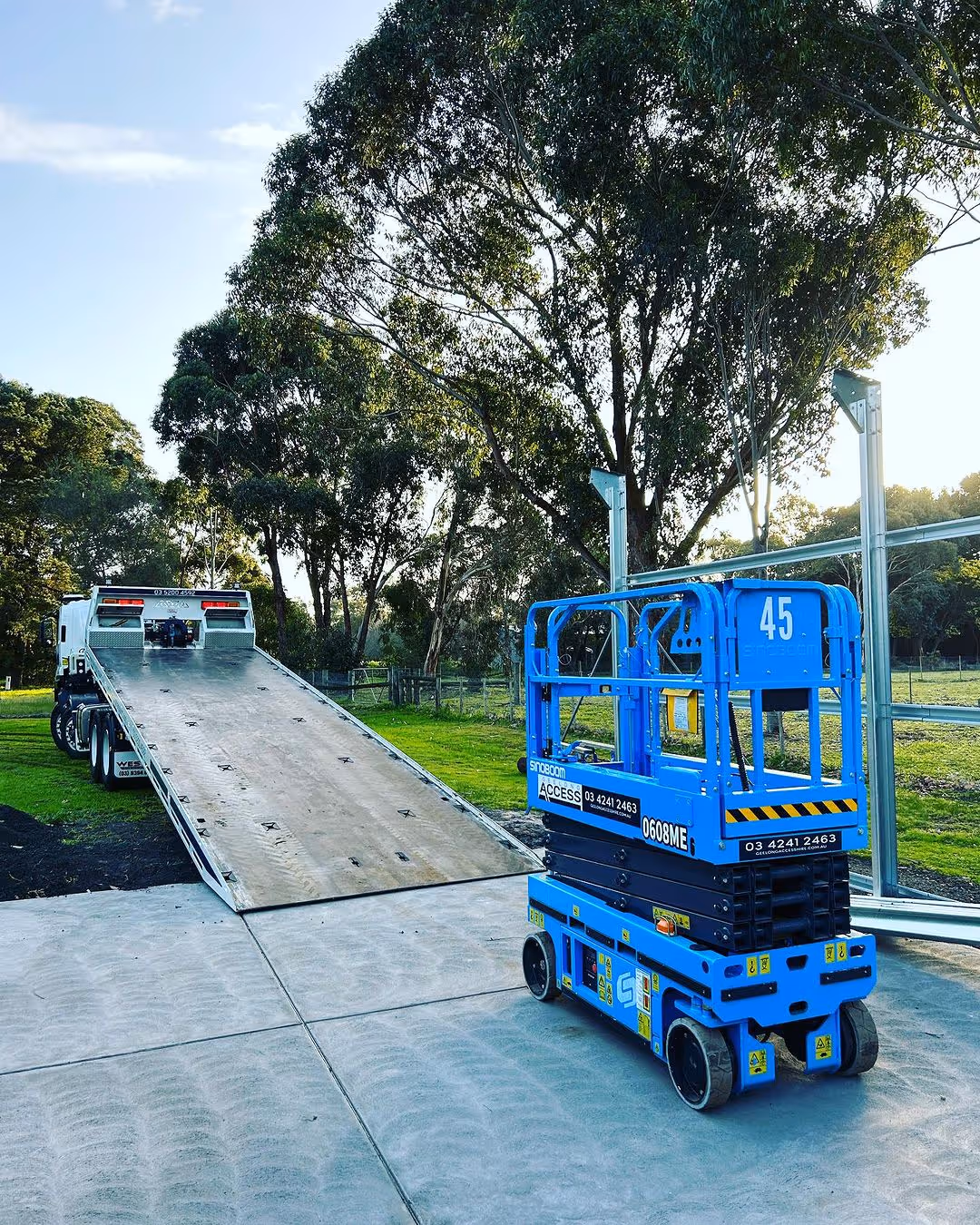 Blue scissor lift on concrete, near an inclined truck ramp, with trees in the background.