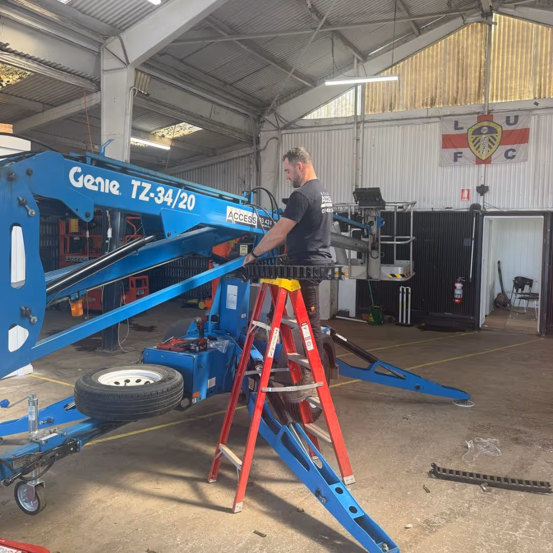 A man stands on a red ladder working on a blue mechanical lift inside a workshop.